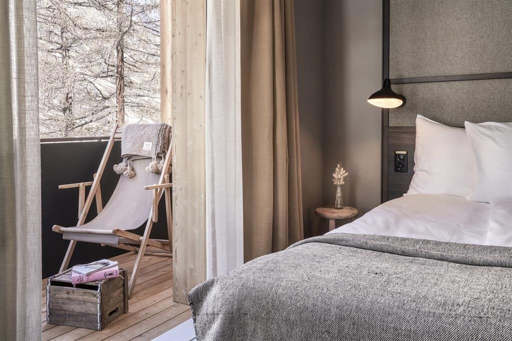 Blick aus dem Hotelzimmer mit Bett auf einen Holzbalkon mit Klappstuhl und Tannen.