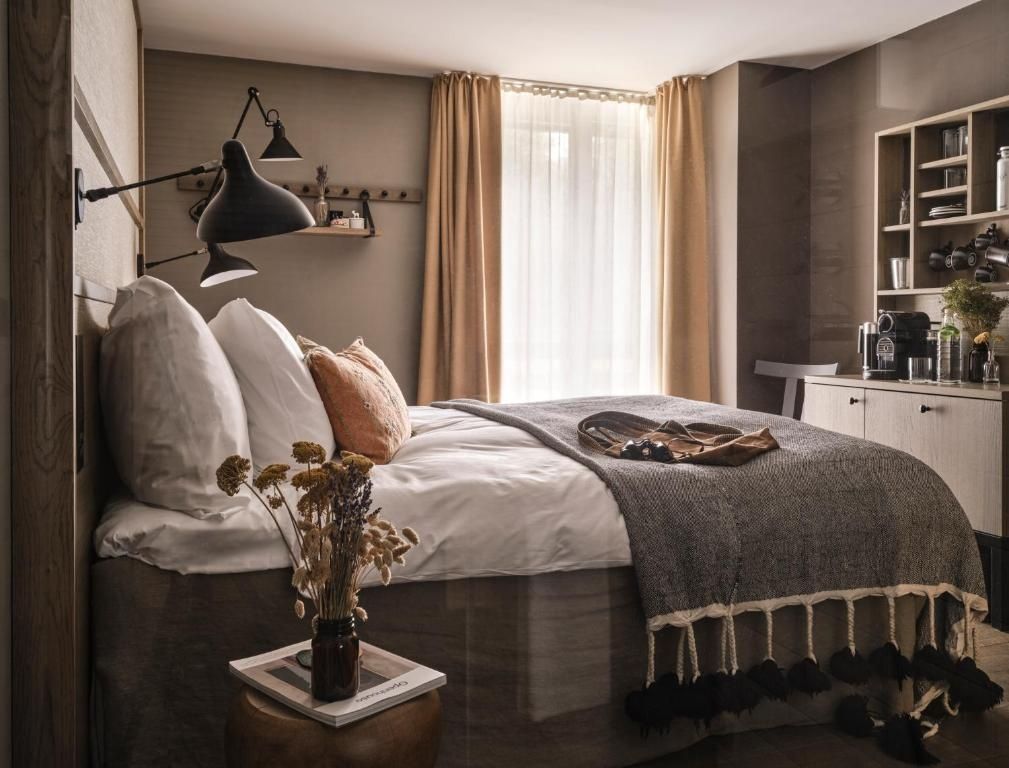 Doppelbett mit grauer Tagesdecke, Kissen und hellen Vorhängen im Hintergrund.