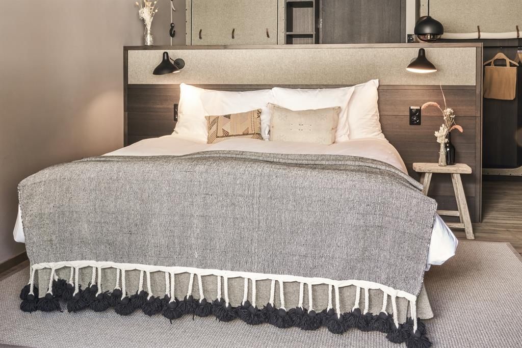 Doppelbett mit grauer Tagesdecke, Fransen und weißen Kissen vor Holzwand.