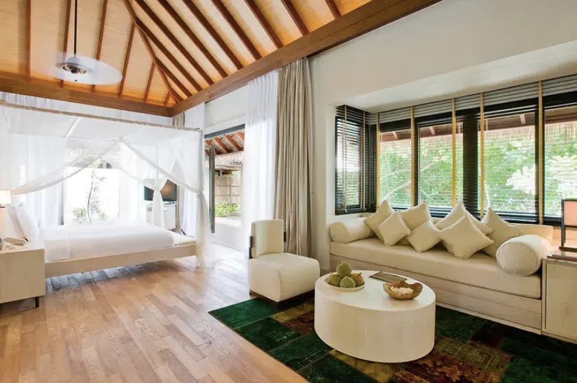 Heller Wohnbereich einer Villa mit Holzboden, weißem Sofa, Reetdach und Blick zum Bett.