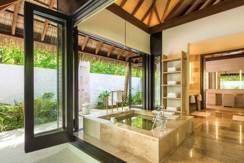 Badezimmer mit großer versenkter Steinbadewanne und geöffneter Tür zum Außenbereich.