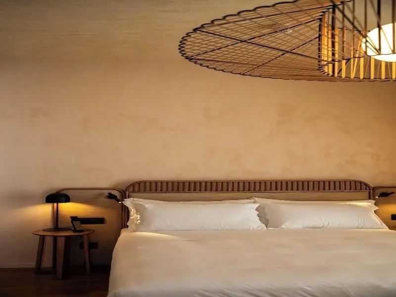 Doppelbett mit weisser Bettwaesche, Holzkopfteil und einer grossen Flechtlampe an der Decke.
