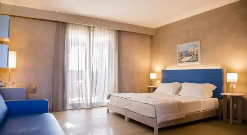 Helles Doppelzimmer mit blauem Kopfteil am Bett und großen Gardinen vor der Fensterfront.