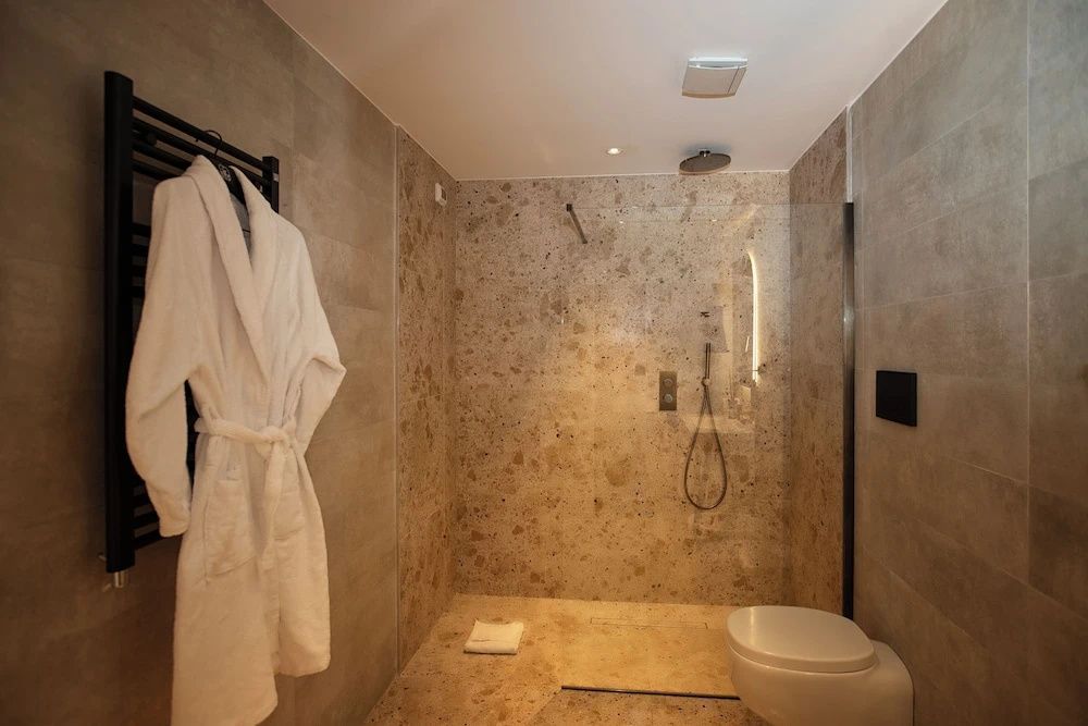Badezimmer mit bodengleicher Dusche, Natursteinfliesen, Bademantel und Toilette.
