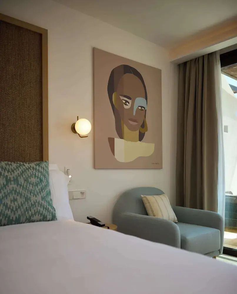Hotelzimmer mit gemaltem Frauenporträt an der Wand, Sessel und Bett mit Kissen.