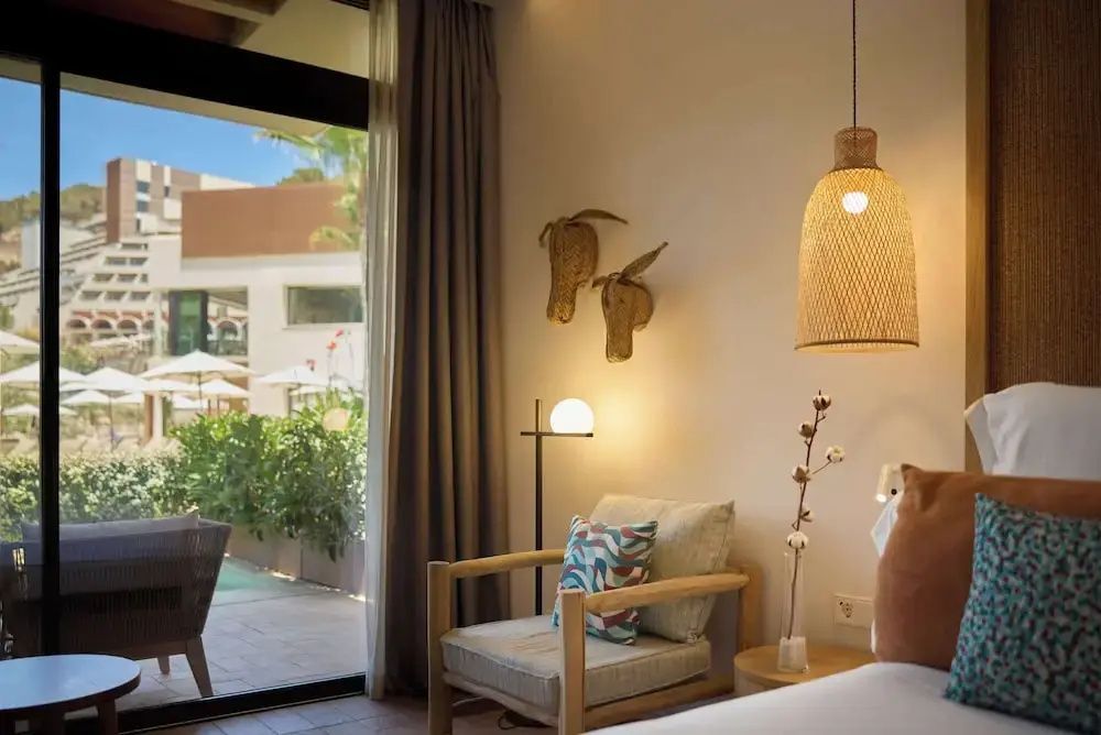 Sitzbereich im Hotelzimmer mit Holzsessel, Hängelampe aus Geflecht und Blick ins Freie.