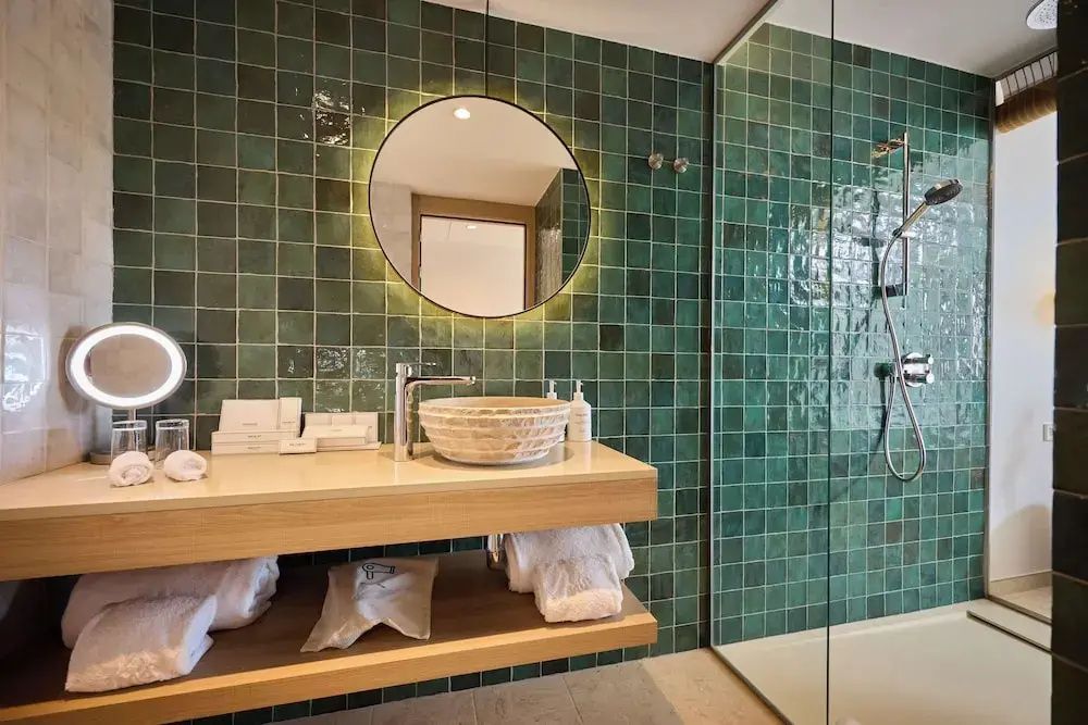 Badezimmer mit grünen Wandfliesen, rundem beleuchtetem Spiegel und Steinwaschbecken.