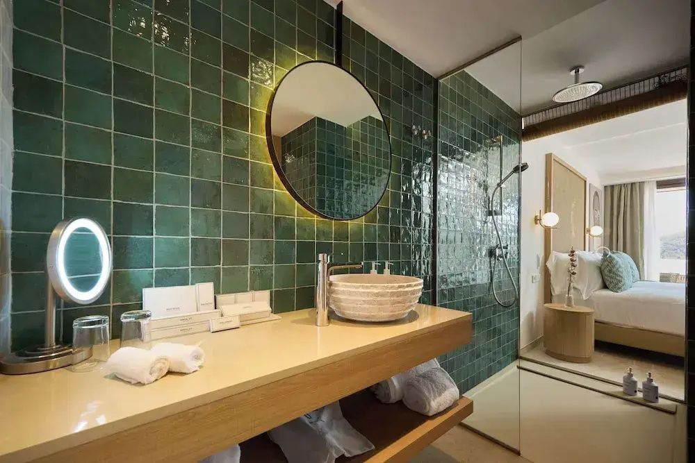 Modernes Bad mit grünen Fliesen, rundem Wandspiegel, Waschtisch und Dusche mit Glaswand.