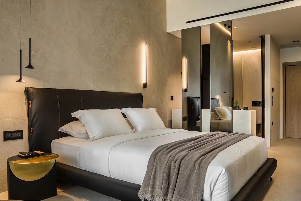 Doppelbett mit weißer Bettwäsche, brauner Tagesdecke und moderner Wandbeleuchtung im Hotelzimmer.