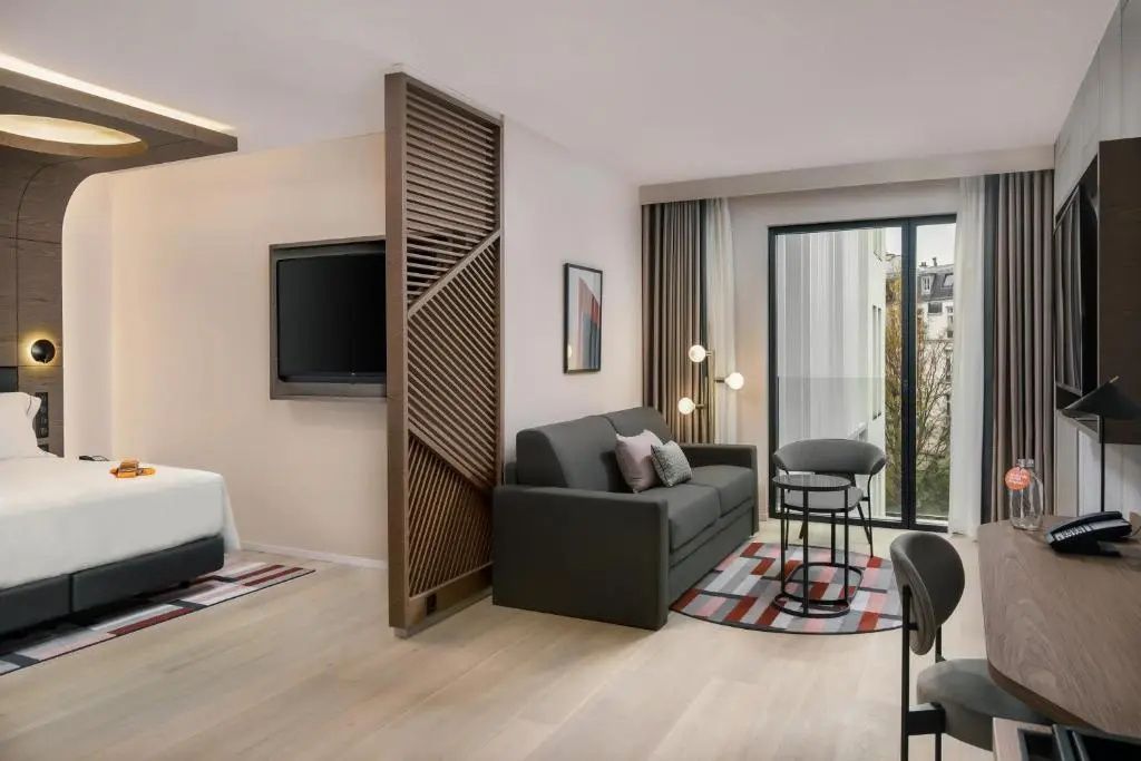 Sitzecke im Zimmer mit dunklem Sofa, Holzraumteiler, Fernseher und Fensterfront.