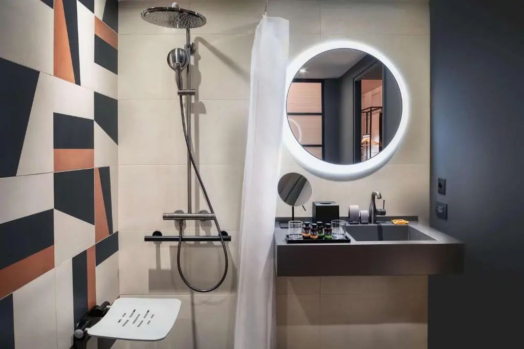 Badezimmer mit Regendusche, klappbarem Duschsitz und rundem beleuchtetem Spiegel über dem Waschtisch.