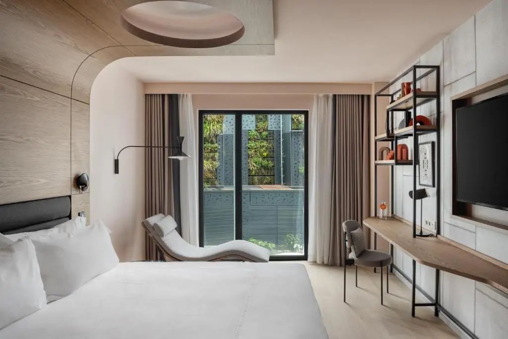 Helles Hotelzimmer mit Liegestuhl, Schreibtisch, Regalelement und Blick zum Fenster.