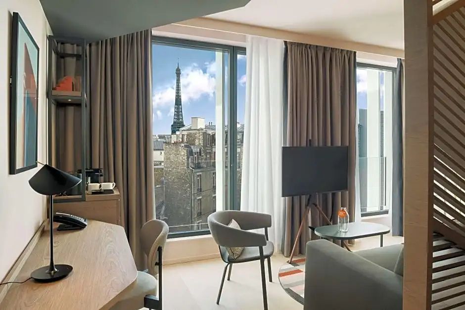 Helles Hotelzimmer mit Sessel, Fernseher auf Stativ und Blick auf den Eiffelturm.