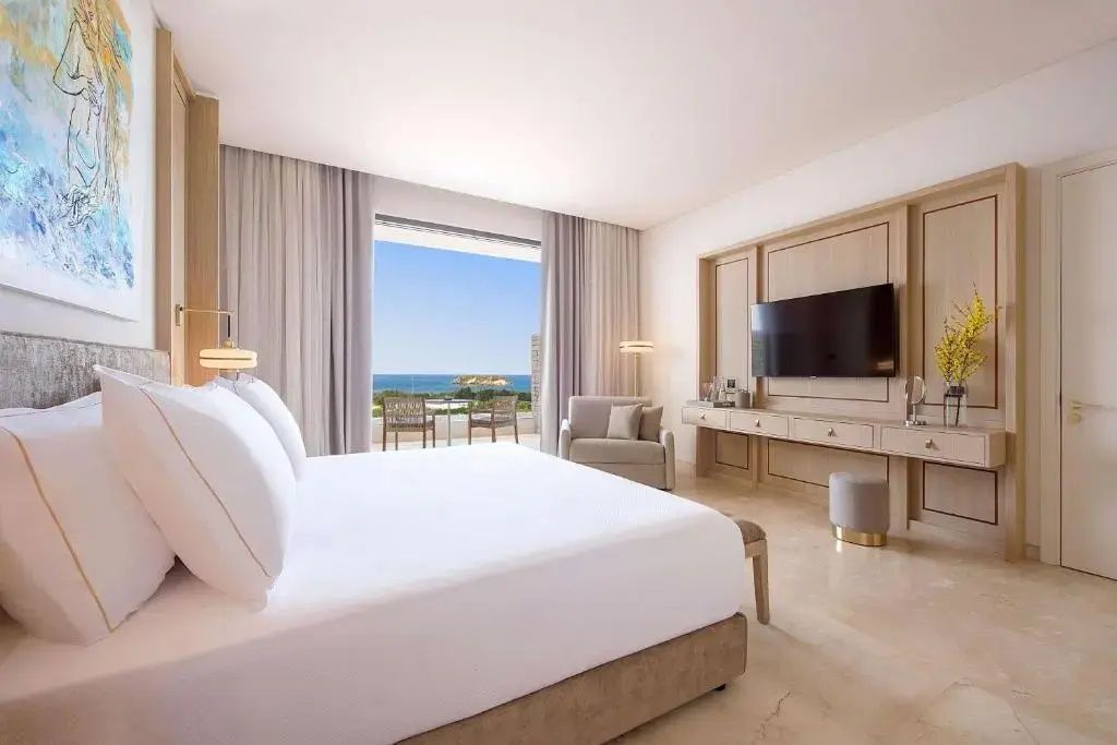 Helles Hotelzimmer mit großem Doppelbett, Wandfernseher und Balkon mit Blick auf das Meer.