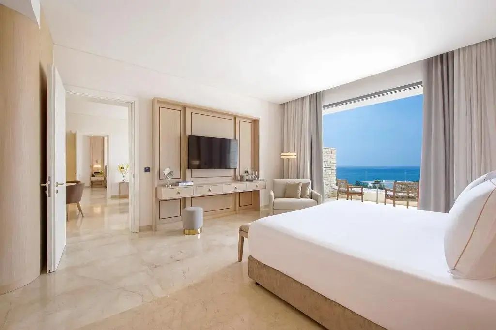 Hell eingerichtetes Hotelzimmer mit großem Doppelbett, Fernseher und Balkon mit Blick auf das Meer.