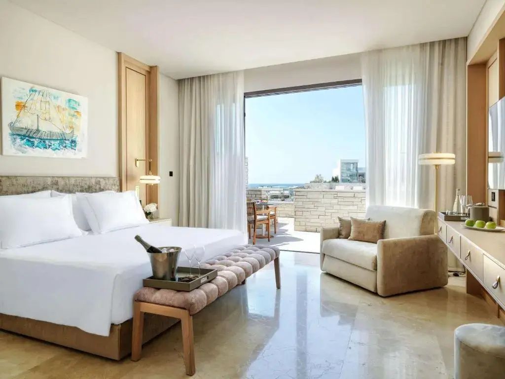 Helles Hotelzimmer mit großem Doppelbett, Sitzbereich und Zugang zur sonnigen Terrasse mit Meerblick.