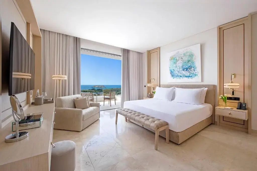 Helles Hotelzimmer mit Doppelbett, beigem Sessel und Balkon mit Blick auf das Meer.