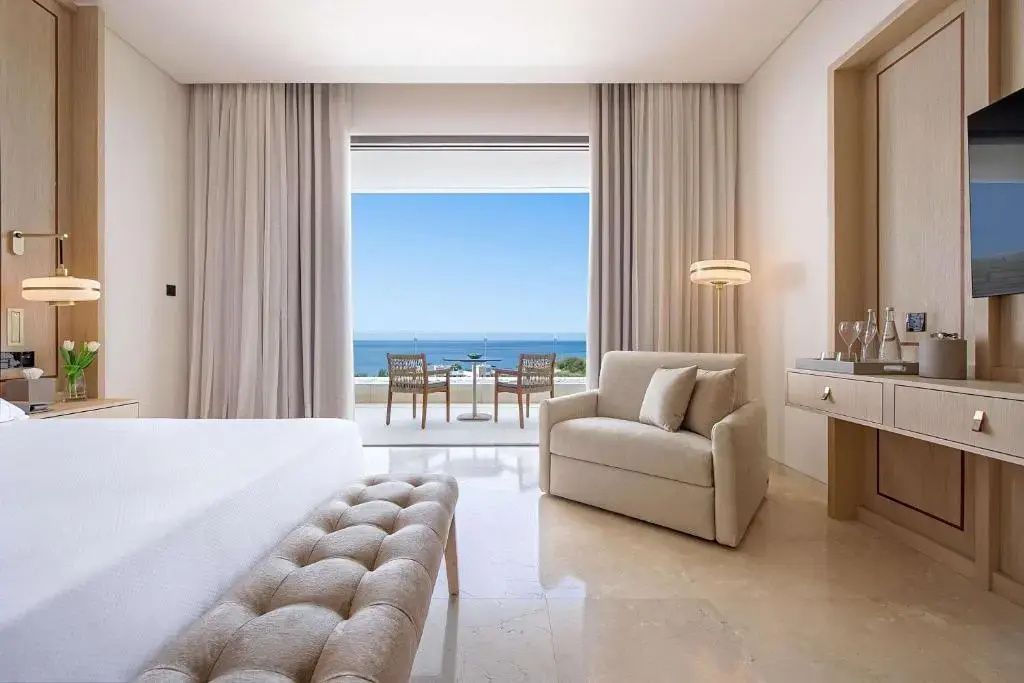 Helles Hotelzimmer mit Sitzgelegenheiten und freiem Blick auf das Meer durch die Balkontür.
