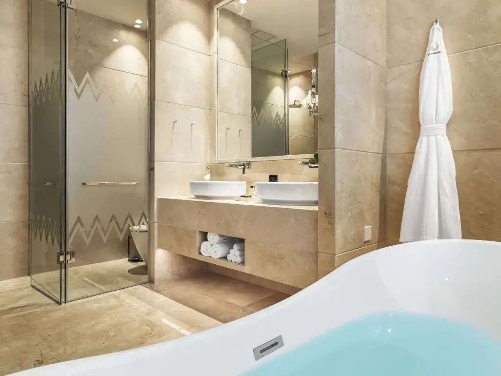 Badezimmer mit beigefarbenen Natursteinfliesen, Doppelwaschtisch, großem Spiegel und Wannenrand im Vordergrund.