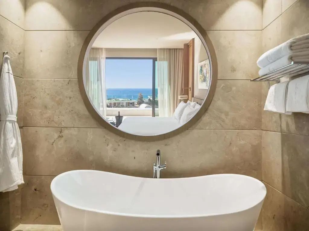 Freistehende weiße Badewanne vor einer Steinwand mit einem großen runden Spiegel.
