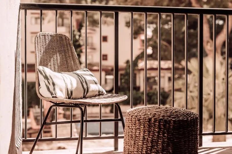Gemütlicher Flechtstuhl mit Kissen und ein Hocker auf einem Balkon hinter einem Geländer