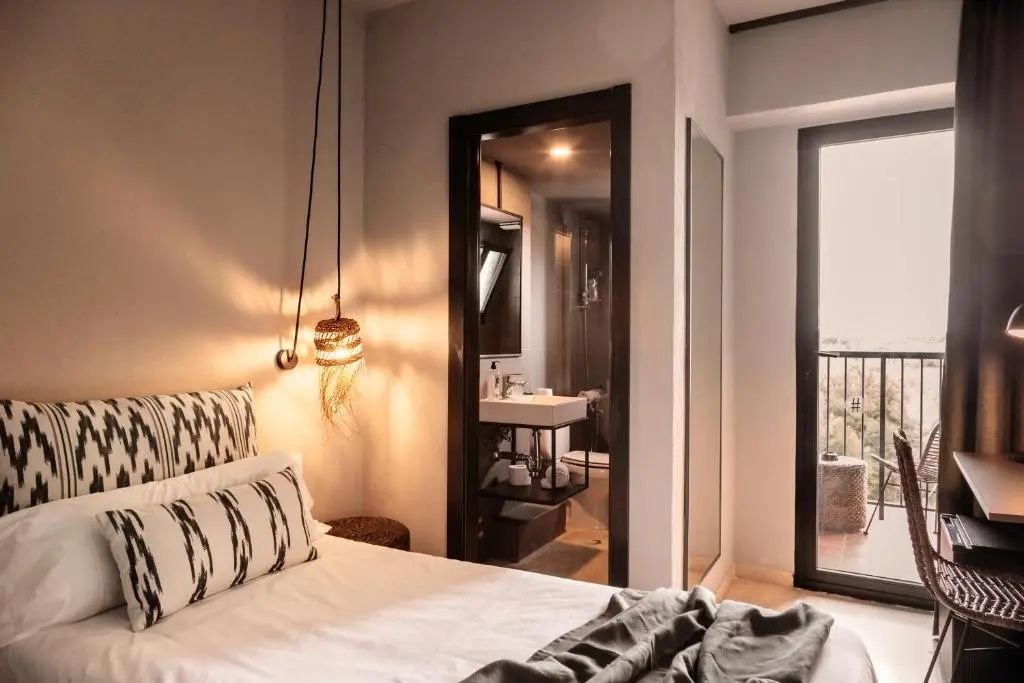 Hell gestaltetes Hotelzimmer mit Bett, Pendelleuchte, Blick ins Badezimmer und Balkonzugang.