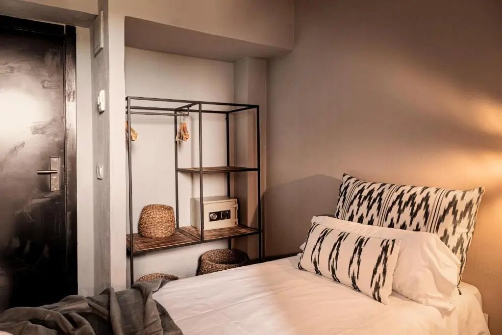 Schlafbereich im Hotelzimmer mit Bett, gemusterten Kissen und offenem schwarzem Regal mit Safe.