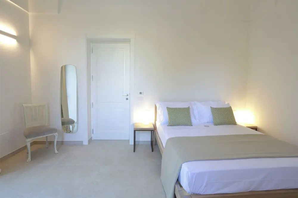 Helles Hotelzimmer mit Doppelbett, weißer Tür, Beistelltisch und einem schmalen Wandspiegel.