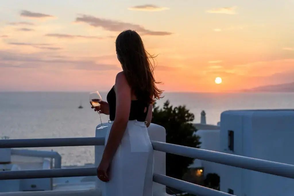 Frau steht abends auf einem Balkon mit Weinglas und blickt auf das Meer.