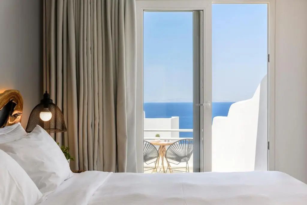 Blick vom Bett durch die geöffnete Balkontür auf das blaue Meer und die Terrasse.