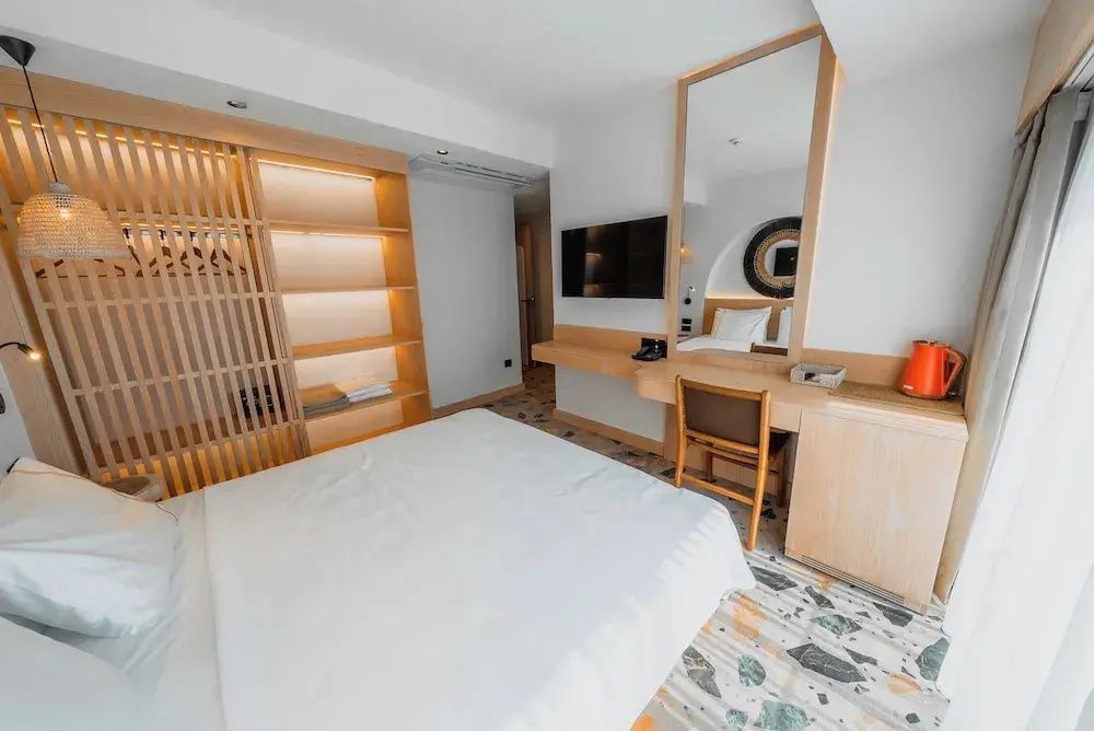 Helles Hotelzimmer mit Doppelbett, Terrazzoboden, Fernseher und offenem Holzkleiderschrank.