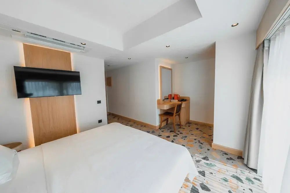 Doppelbett in einem hellen Zimmer mit Terrazzoboden und Schreibtischecke im Hintergrund.
