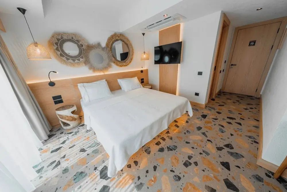Helles Doppelbett mit Holzkopfteil, runden Spiegeln an der Wand und buntem Terrazzoboden.
