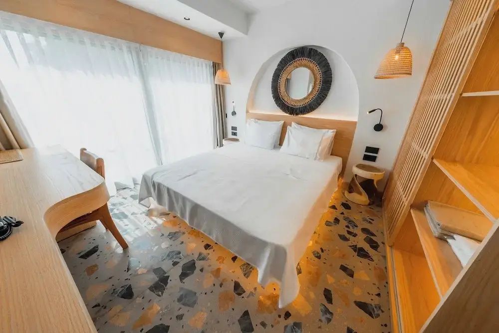 Lichtdurchflutetes Hotelzimmer mit Doppelbett, bodentiefen Vorhängen und buntem Terrazzoboden.