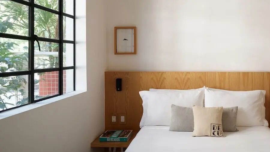 Doppelbett mit holzverkleidetem Kopfteil, weißen Kissen und Fenster mit schwarzem Rahmen.