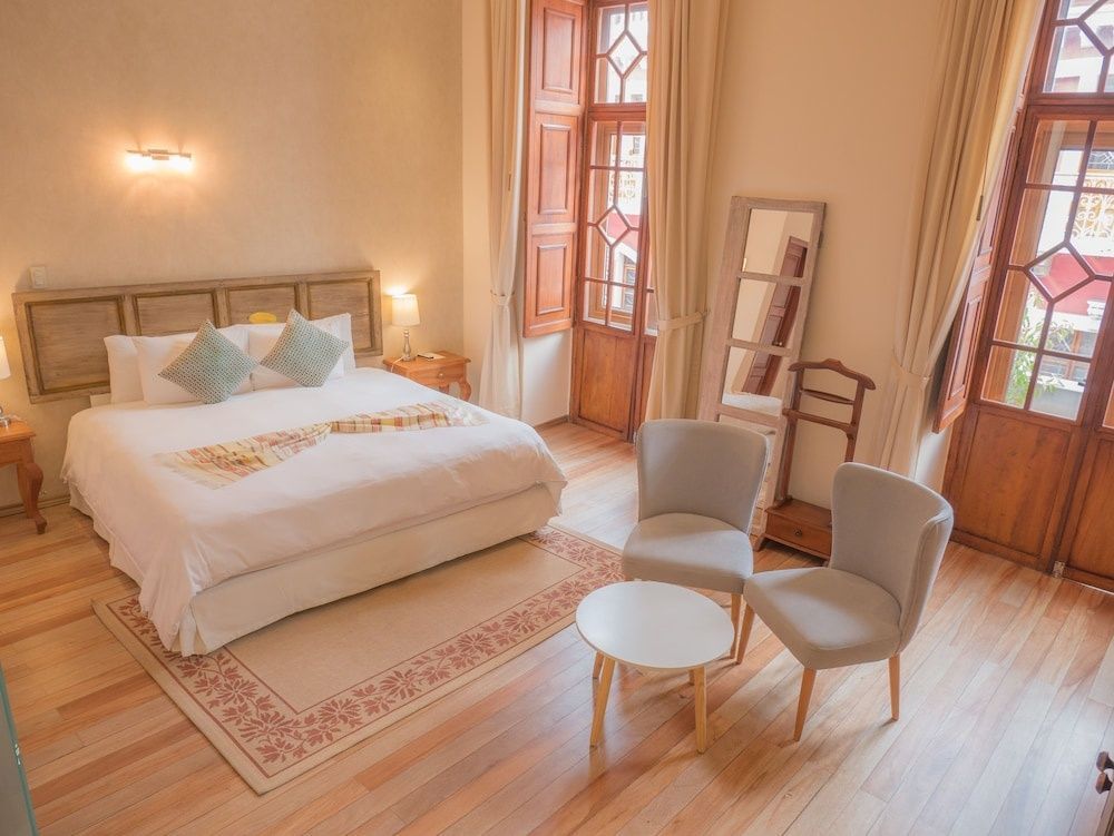 Helles Hotelzimmer mit Doppelbett, Sitzbereich mit zwei Sesseln und hohen Holzfenstern.