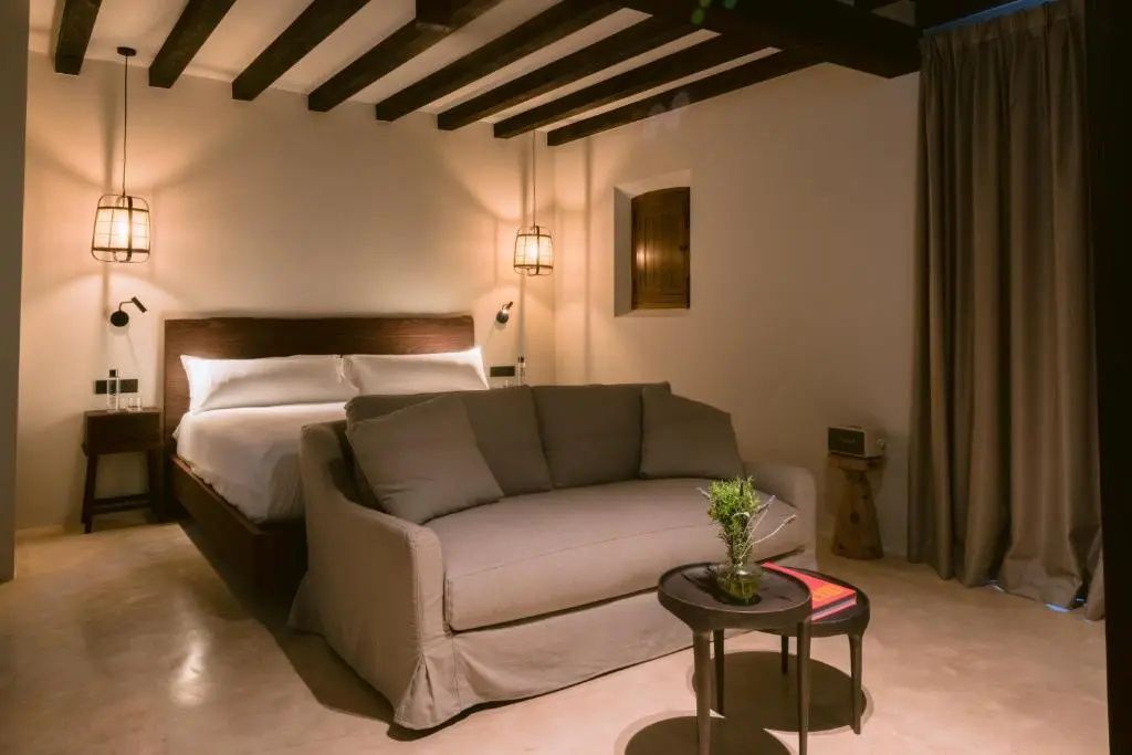 Hotelzimmer mit dunkler Holzbalkendecke, weißem Doppelbett und beigem Sofa.