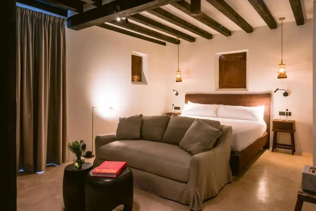 Schlafzimmer mit weiß eingedecktem Doppelbett, beigem Sofa und dunklen Holzbalken.