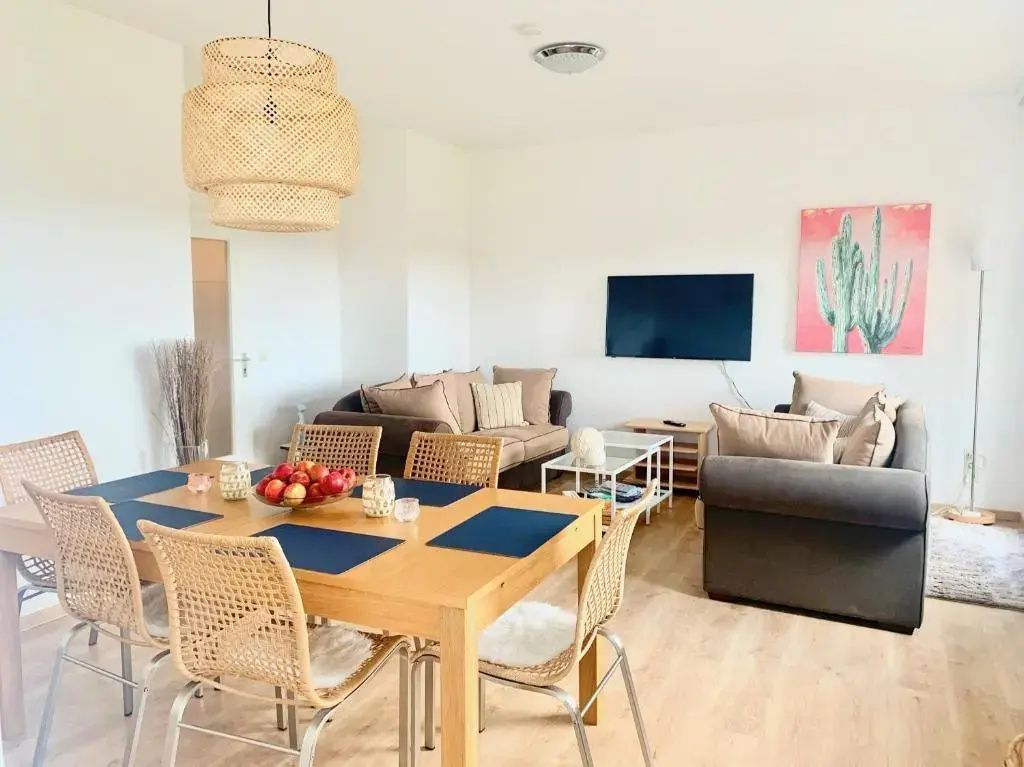Helles Wohnzimmer mit Holztisch, Flechtstühlen, braunen Sofas und einem Fernseher an der Wand.