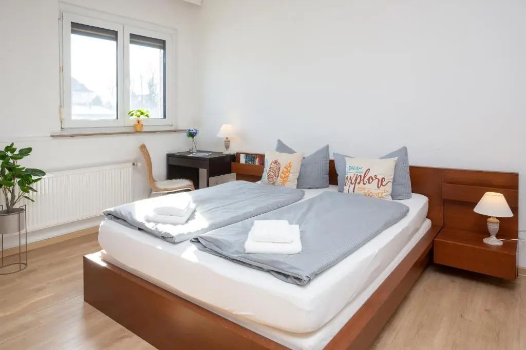 Schlafbereich mit Doppelbett aus Holz, grauer Bettwäsche und Schreibtisch am Fenster.