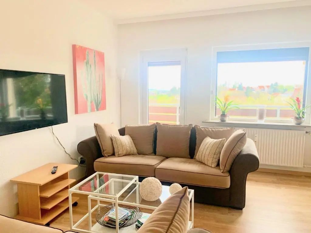 Wohnzimmer mit braunem Sofa vor Fenstern, Glastisch und Wandbild mit Kaktusmotiv.