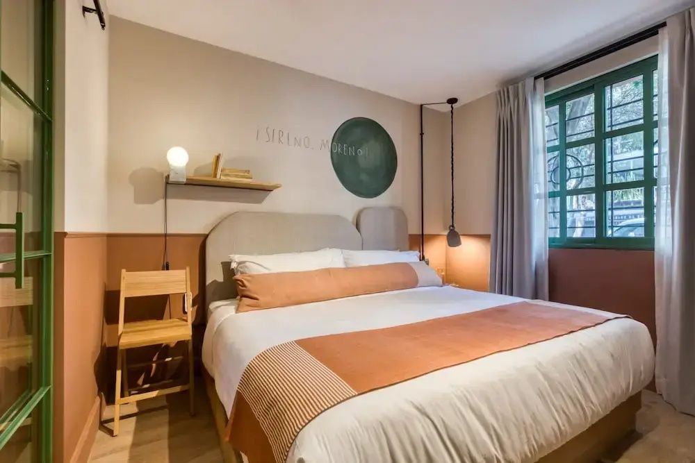 Doppelbett mit orangefarbener Kissenrolle in einem hellen Zimmer mit Stuhl und Fenster.
