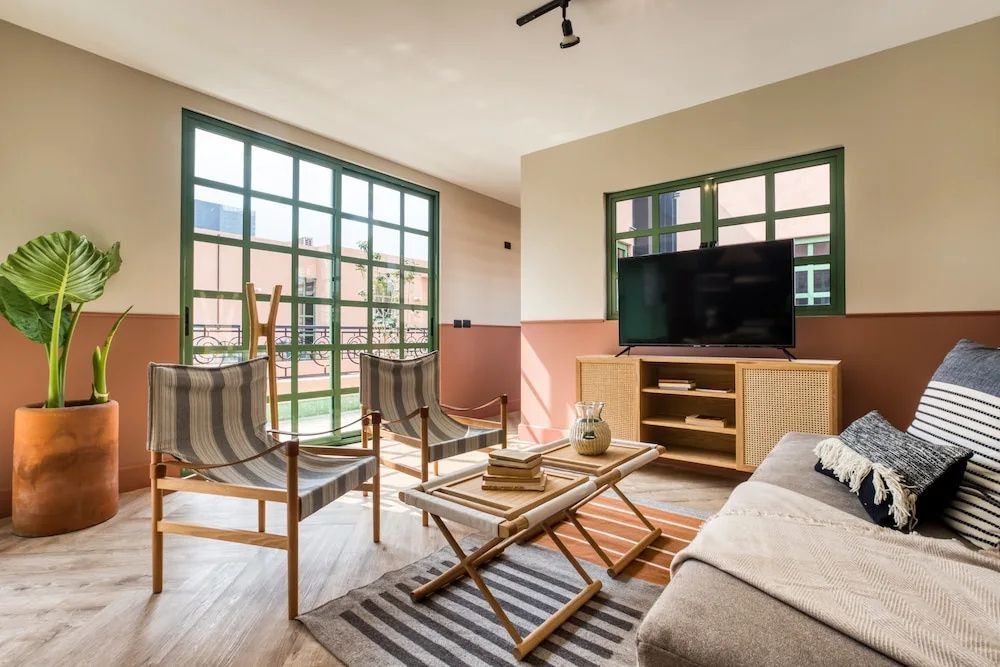Wohnbereich mit Fernseher auf Holzkommode, zwei Streifenstühlen und großen grünen Fenstern.