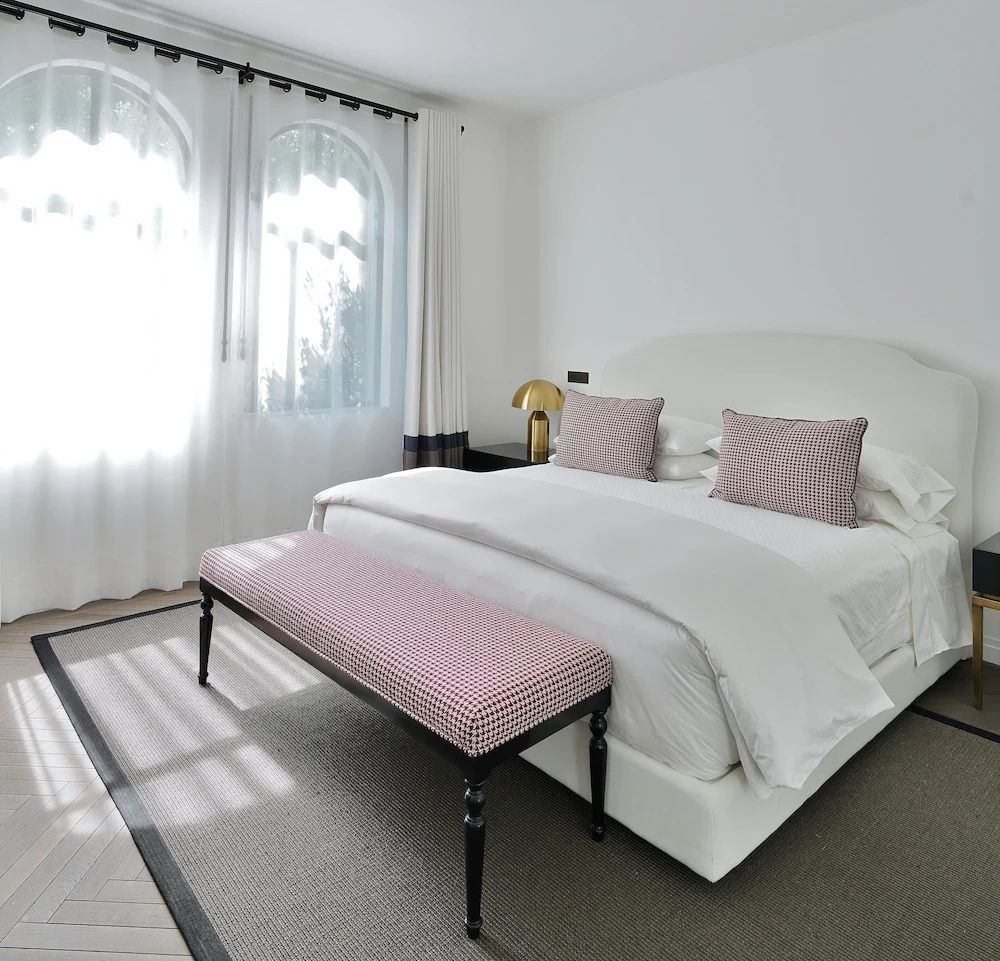 Hell eingerichtetes Schlafzimmer mit weißem Doppelbett, Bank und großen Rundbogenfenstern.