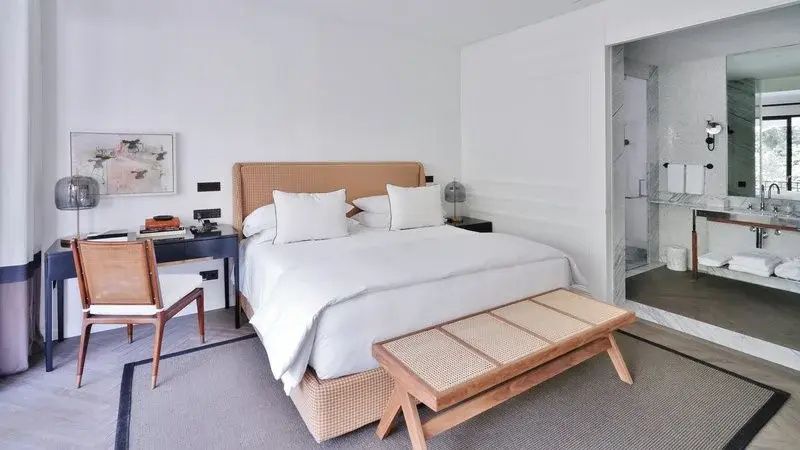 Doppelbett mit weißer Bettwäsche, geflochtenem Kopfteil und Holzbank davor im hellen Hotelzimmer.