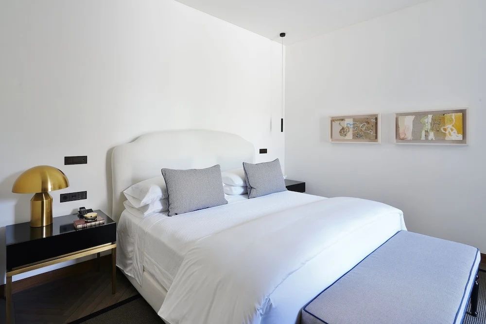 Weißes Doppelbett mit grauen Kissen und gepolsterter Sitzbank in hellem Hotelzimmer.