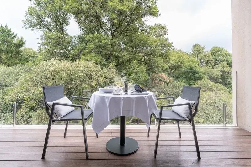 Gedeckter Tisch mit zwei Stühlen auf einer Terrasse mit Blick auf grüne Bäume.