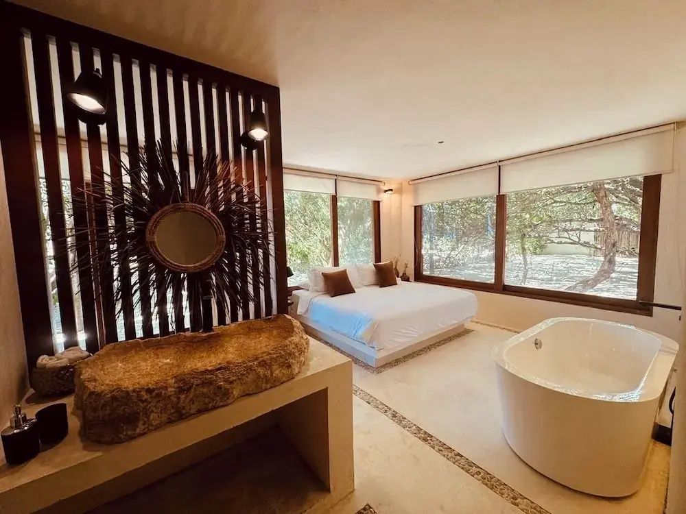 Helles Hotelzimmer mit Doppelbett, großer Badewanne, Natursteinwaschbecken und Panoramafenstern.