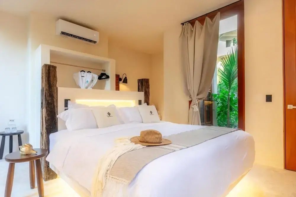 Hell eingerichtetes Hotelzimmer mit Doppelbett, Strohhut auf der Bettdecke und Balkontür ins Grüne.