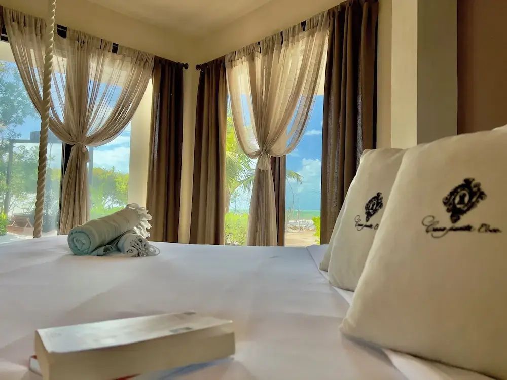 Blick über ein weiß bezogenes Doppelbett mit Kissen auf große Fenster mit Vorhängen.